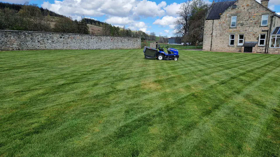 Ride-on mower maintaining a broad striped lawn beside a stone property